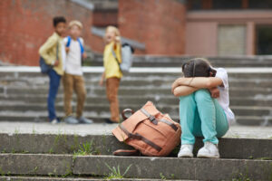 three children hazing another child who is sitting on the ground with their folded arms covering their head.