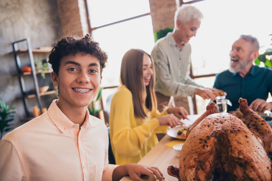 a young person with braces n the foreground smiling while people in the backgroun eating, Holiday Braces & Aligners Care: Protect Your Smile During Thanksgiving Feasts